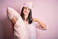 Young brunette woman with blue eyes wearing pajama hugging bed pillow at night time with surprise face pointing finger to himself