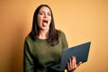 Young brunette woman with blue eyes working using computer laptop over yellow background sticking tongue out happy with funny Royalty Free Stock Photo