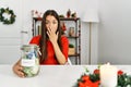 Young brunette woman on christmas holding charity jar with money at home covering mouth with hand, shocked and afraid for mistake Royalty Free Stock Photo