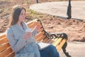 Young brunette woman in coat sits on bench in city park, eats bun with sugar and drinks coffee. Springtime. Royalty Free Stock Photo