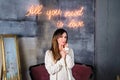 Young brunette woman in a cozy sweater standing in front of All you need is love neon sign on background