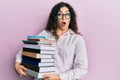 Young brunette woman with curly hair holding a pile of books afraid and shocked with surprise and amazed expression, fear and Royalty Free Stock Photo