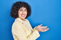 Young brunette woman with curly hair standing over blue background pointing aside with hands open palms showing copy space, Royalty Free Stock Photo