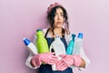 Young brunette woman with curly hair wearing cleaner apron holding cleaning products in shock face, looking skeptical and Royalty Free Stock Photo