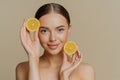 Young brunette woman with dark combed hair looks directly at camera holds two slices of lemons stands topless has