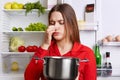 Young brunette woman with displeased expression smells spoiled soup in stew pan, feels musty smell at home kitchen, stands against