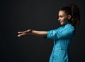 Young brunette woman doctor nurse in blue medical gown holds out her hands with open palms with copy space. Side view