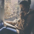 Young brunette woman drinking a cup of tea or coffee and reading a book. Female sitting at home by the window and read a book Royalty Free Stock Photo