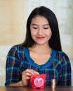 Young brunette woman facing camera, holding piggy bank and placing money inside it, smiling happily Royalty Free Stock Photo