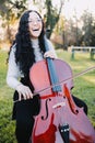 Happy young brunette woman with glasses laughing and playing cello at sunset in the park. Vertical Royalty Free Stock Photo