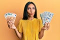 Young brunette woman holding bunch of dollars and euros making fish face with mouth and squinting eyes, crazy and comical Royalty Free Stock Photo