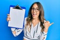 Young brunette woman holding clipboard with blank space pointing thumb up to the side smiling happy with open mouth Royalty Free Stock Photo