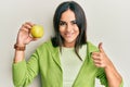 Young brunette woman holding green apple smiling happy and positive, thumb up doing excellent and approval sign