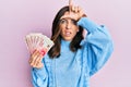 Young brunette woman holding 20 israel shekels banknotes making fun of people with fingers on forehead doing loser gesture mocking