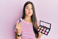 Young brunette woman holding makeup brush and blush palette puffing cheeks with funny face