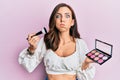 Young brunette woman holding makeup brush and blush puffing cheeks with funny face