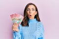 Young brunette woman holding 100 new zealand dollars banknote scared and amazed with open mouth for surprise, disbelief face Royalty Free Stock Photo