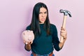 Young brunette woman holding piggy bank and hammer clueless and confused expression Royalty Free Stock Photo