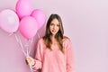 Young brunette woman holding pink balloons thinking attitude and sober expression looking self confident