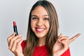 Young brunette woman holding red lipstick smiling happy pointing with hand and finger to the side Royalty Free Stock Photo