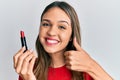 Young brunette woman holding red lipstick smiling happy and positive, thumb up doing excellent and approval sign Royalty Free Stock Photo