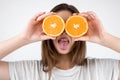 Young brunette woman holding two halves of oranges near her eyes showing tongue standing on isolated white background dietology