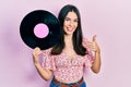 Young brunette woman holding vinyl disc smiling happy and positive, thumb up doing excellent and approval sign Royalty Free Stock Photo