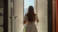 Young brunette woman in long dress opens balcony doors, walks out on the terrace and enjoys sunrise or sunset over the Royalty Free Stock Photo