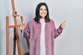 Young brunette woman painter sitting at art studio holding art word smiling happy and positive, thumb up doing excellent and Royalty Free Stock Photo