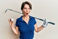 Young brunette woman with short hair holding ball and golf club afraid and shocked with surprise and amazed expression, fear and Royalty Free Stock Photo