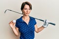 Young brunette woman with short hair holding ball and golf club relaxed with serious expression on face