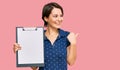 Young brunette woman with short hair holding clipboard with blank space pointing thumb up to the side smiling happy with open Royalty Free Stock Photo