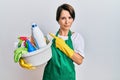 Young brunette woman with short hair wearing apron holding cleaning products pointing aside worried and nervous with forefinger,