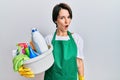 Young brunette woman with short hair wearing apron holding cleaning products in shock face, looking skeptical and sarcastic, Royalty Free Stock Photo