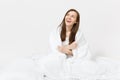 Young brunette woman sitting in bed with white sheet, pillow, wrapping in blanket on white background. Beauty female