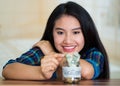 Young brunette woman sitting down facing camera, putting coins inside glass jar with money inside, label reading