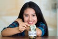 Young brunette woman sitting down facing camera, putting coins inside glass jar with money inside, label reading