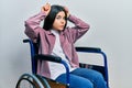 Young brunette woman sitting on wheelchair doing funny gesture with finger over head as bull horns