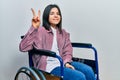 Young brunette woman sitting on wheelchair smiling looking to the camera showing fingers doing victory sign Royalty Free Stock Photo