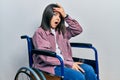 Young brunette woman sitting on wheelchair surprised with hand on head for mistake, remember error