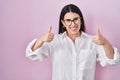 Young brunette woman standing over pink background success sign doing positive gesture with hand, thumbs up smiling and happy Royalty Free Stock Photo