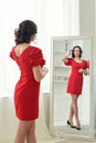 Young brunette woman stands looking in reflection in a full-length mirror in a short red dress in heels