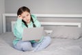 young brunette woman with two pigtails in turquoise hoodie and jeans sitting on a bed with white sheets with laptop