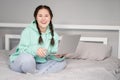 young brunette woman with two pigtails in turquoise hoodie and jeans sitting on a bed with white sheets with laptop