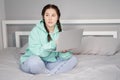 young brunette woman with two pigtails in turquoise hoodie and jeans sitting on a bed with white sheets with laptop