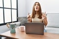 Young brunette woman using laptop at home drinking a cup of coffee amazed and surprised looking up and pointing with fingers and Royalty Free Stock Photo