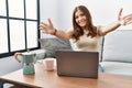 Young brunette woman using laptop at home drinking a cup of coffee looking at the camera smiling with open arms for hug Royalty Free Stock Photo