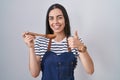 Young brunette woman wearing apron tasting food holding wooden spoon smiling happy and positive, thumb up doing excellent and