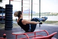 Young brunette woman, wearing black fitness overall, doing abs on horizontal bars on sports playground by city lake in summer. Fit