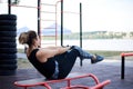 Young brunette woman, wearing black fitness overall, doing abs on horizontal bars on sports playground by city lake in summer. Fit